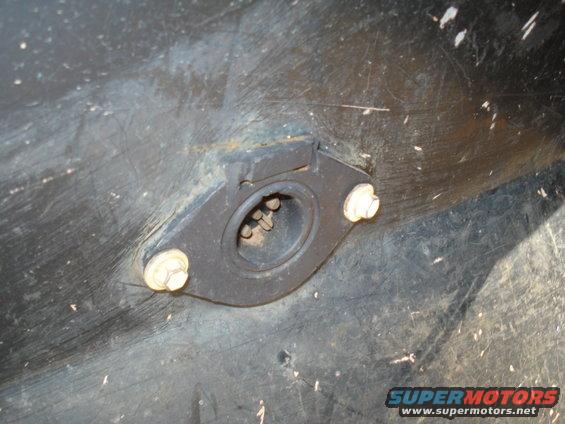 Close-up of a black plastic part with a metal flange and two bolts holding it in place. A circular opening in the flange shows some metal prongs