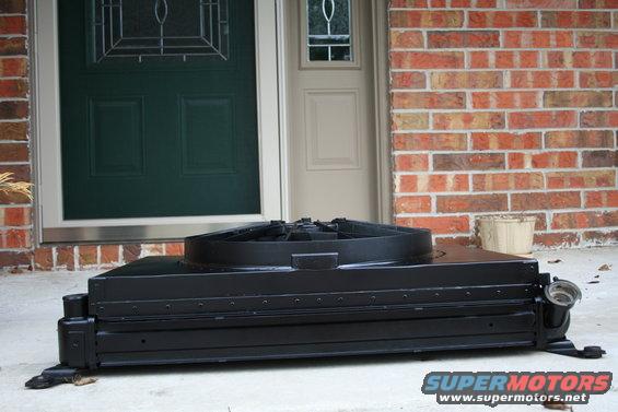 Black radiator fan shroud with fan, resting on concrete in front of a brick wall and double doors