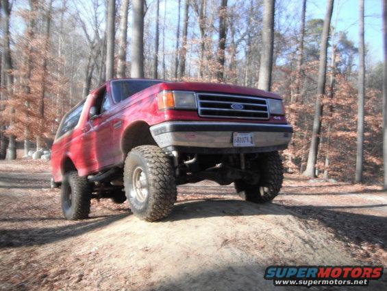 Red 1989 Ford Bronco with lifted suspension driving on a dirt mound in a wooded area