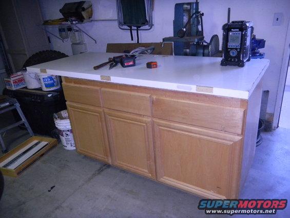 Workbench with cabinets, clamp, tape measure, and radio on top. Tools and buckets in background