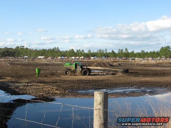 Green monster truck with large tires driving through mud, with spectators and tents in the background