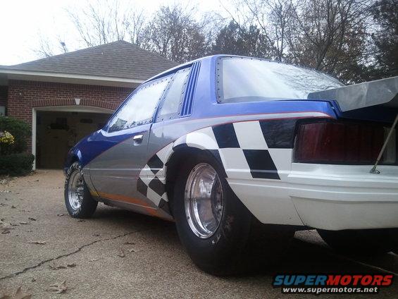 Customized Ford Mustang drag car with checkerboard graphics and a large rear spoiler, parked in a driveway
