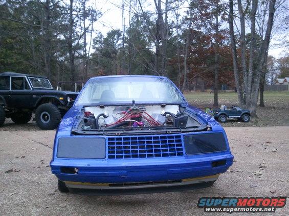 Blue car with hood open, showing engine bay. Black Ford Bronco in background