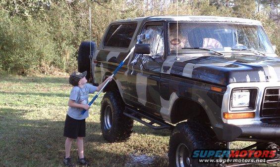 Child washes a lifted, camouflage-painted Ford Bronco with a brush on a pole