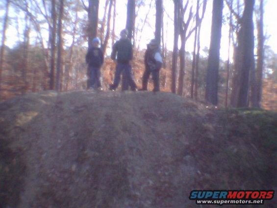 Three people stand on a dirt mound in a wooded area