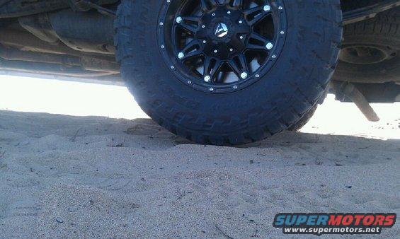Close-up of a black aftermarket wheel and tire on sand, showing part of the vehicle's undercarriage