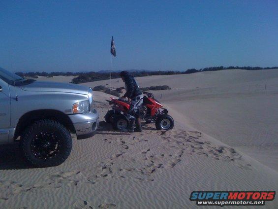 Silver pickup truck parked next to a red ATV with a person on it in sand dunes. A flag is visible in the background