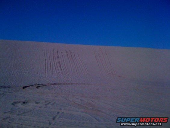 Sand dunes with tire tracks under a clear blue sky. Supermotors.net logo in lower right