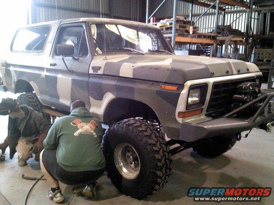 Camouflaged Ford Bronco with large off-road tires and custom bumper, being worked on by two people