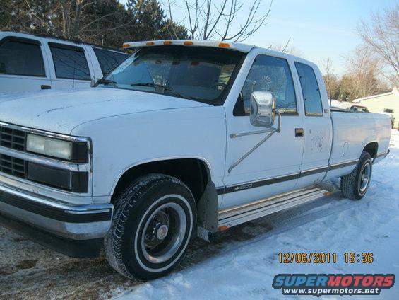 White four-door pickup truck with a visor and running boards, parked in the snow. Another white vehicle is partially visible to the left