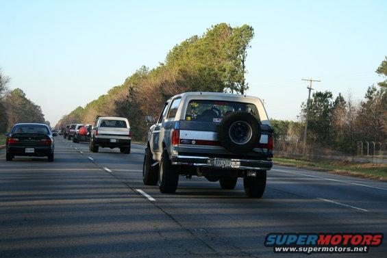 Rear view of a blue and white Ford Bronco driving on a highway. Spare tire mounted on the back. Other cars in the background