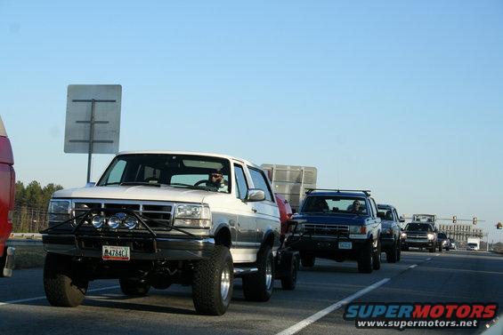 White Ford Bronco with bull bar and off-road lights, towing a trailer, driving on highway with other vehicles