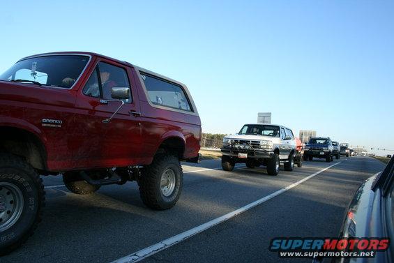 Red Ford Bronco with lifted suspension and large tires, followed by white and dark SUVs on a highway
