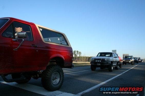 Red Ford Bronco in foreground, followed by white Ford F-150, dark SUV, and other vehicles on highway