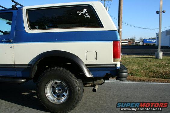Rear quarter view of a blue and white Ford Bronco with a roof rack and a skull and crossbones sticker on the rear window