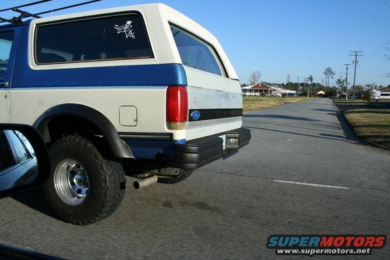 Rear view of a white and blue Ford Bronco driving down a street. It has a skull sticker on the rear window