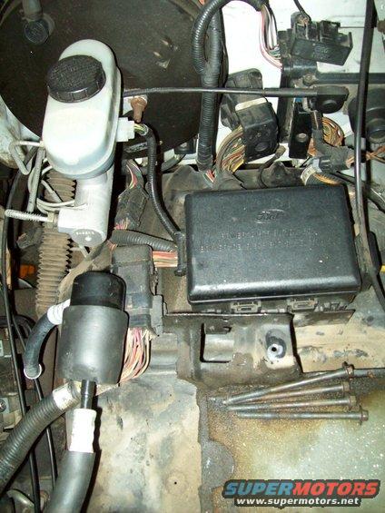 Close-up of engine bay with fuse box, brake fluid reservoir, and wiring harnesses. Ford logo visible on fuse box