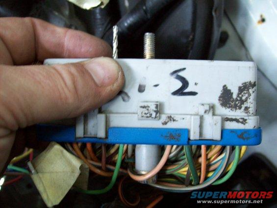 Hand holding a wire near a white automotive electrical connector with blue plastic housing and many colored wires