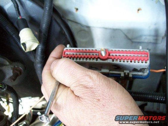Hand holding a gray and red wiring harness connector with a small screwdriver. Black wire loom is visible