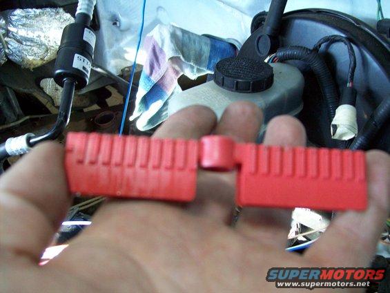 Hand holding a red plastic part in front of engine bay components like brake fluid reservoir and hoses
