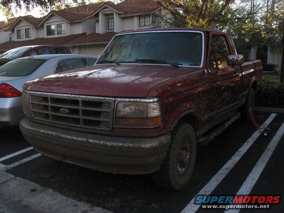 1995 Ford F-150 pickup truck, front view, covered in mud. Another car is parked to the left