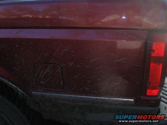 Mud-splattered red pickup truck rear side with fuel door and taillight visible. Supermotors.net logo at bottom right