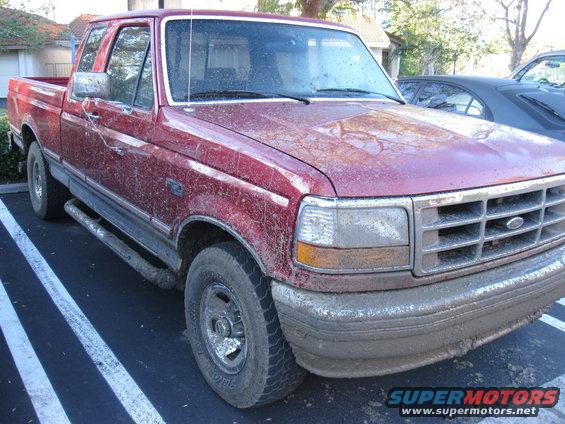 Red pickup truck covered in mud, parked in a lot. Front 3/4 view