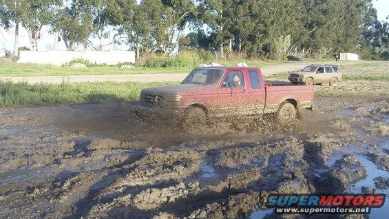 Red pickup truck splashing mud, another SUV behind it on a dirt road