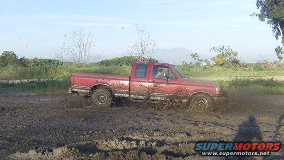 Red pickup truck with a crew cab driving through mud, splashing dirt. The truck appears to be from the early to mid-1990s