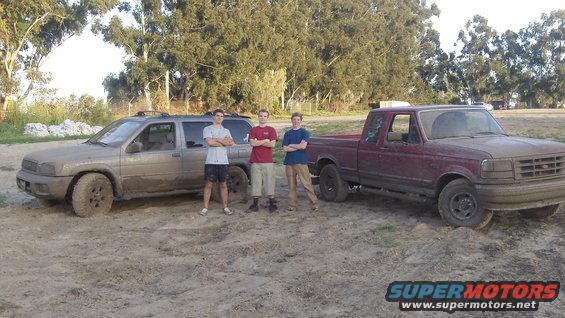 Maroon Ford F-150 pickup truck and a silver SUV covered in mud, parked side-by-side outdoors with three young men standing between them