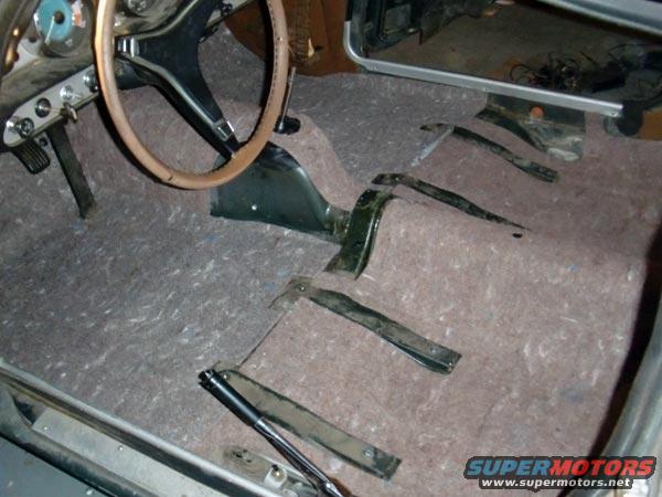Car interior with floor carpet removed, showing pedals, steering wheel, and disassembled center console parts