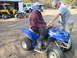 2003 Yamaha RAPTOR 660 - A person sits on a blue Yamaha ATV while an adult looks on. Other vehicles are parked in the sandy area.