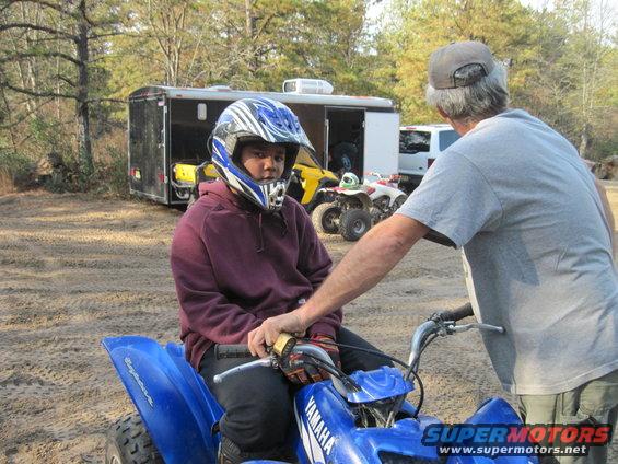 Blue Yamaha ATV with rider in helmet, man standing, trailer and yellow ATV in background