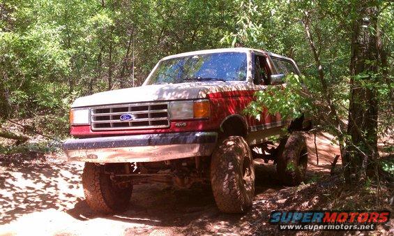 Red and tan 1987 Ford Bronco with lifted suspension and large tires on a dirt trail