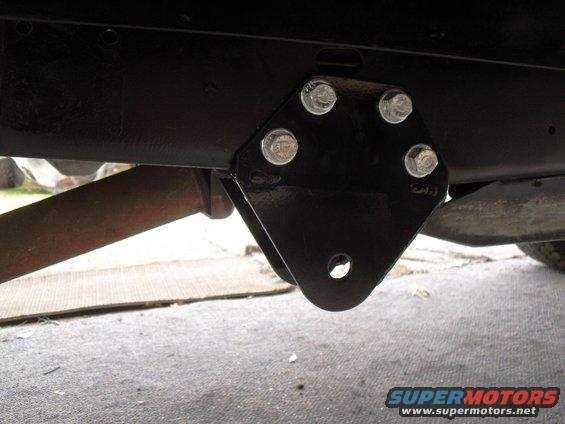 Close-up of a black metal suspension bracket bolted to a vehicle frame, with a rusty pipe visible underneath
