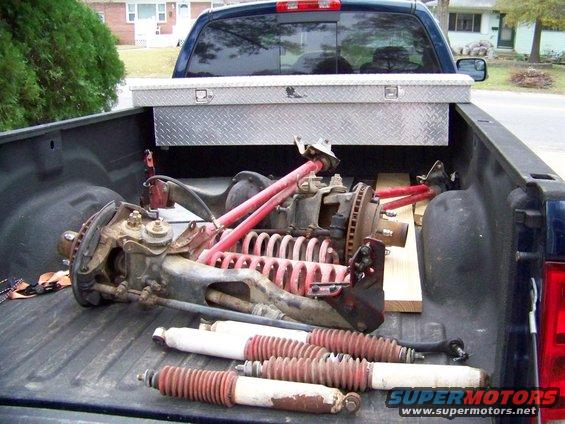 Truck bed loaded with front suspension components, including springs, shocks, and axles. A toolbox is mounted above the parts