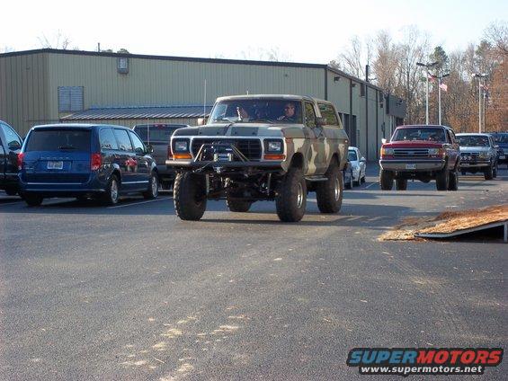 Modified Ford Bronco with camouflage paint and large tires, front view. Other vehicles and a building are in the background