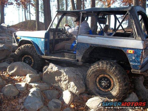Blue and white Ford Bronco off-roading over rocks, driver visible inside