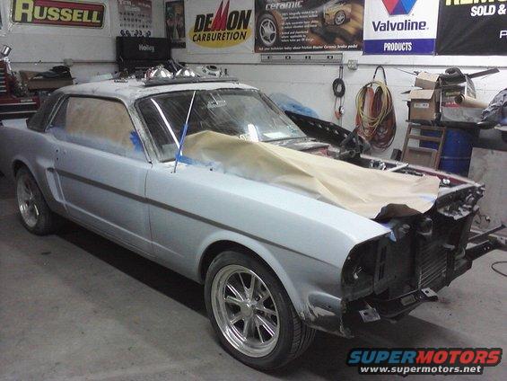 1965 Ford Mustang coupe in primer, hood off, in a garage with "SuperMotors.net" watermark