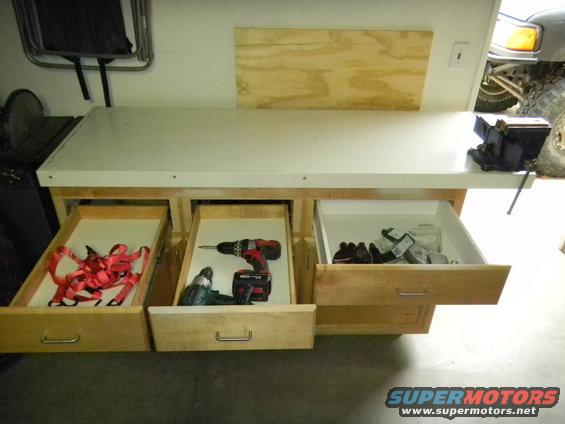 Workbench with drawers open showing red straps, drills, and other tools. A vehicle is partially visible in the background