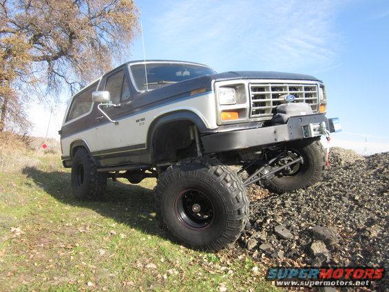 Lifted Ford Bronco with aftermarket bumper and large tires, parked on a grassy slope next to a pile of rocks