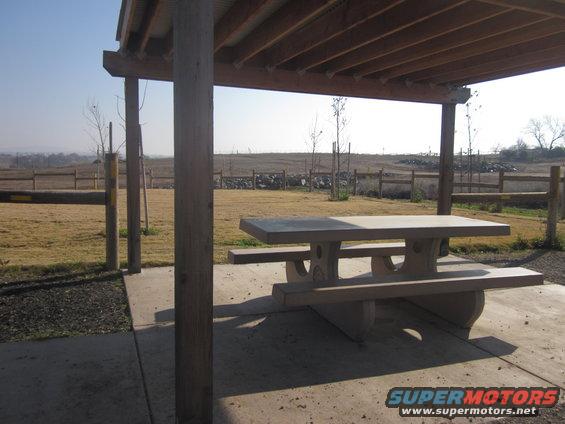 Picnic table under wooden shelter with dry grass and fence in background