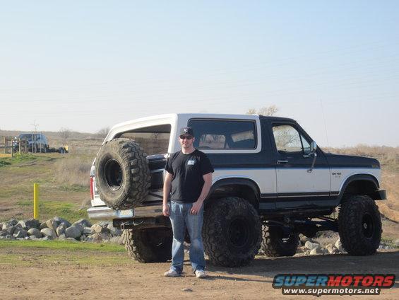 Man standing next to a lifted 1985 Ford Bronco with a spare tire mounted on the rear. The Bronco is white and dark blue