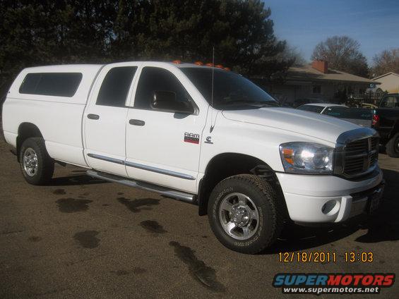 White pickup truck with a camper shell, chrome wheels, and side steps. The truck has a red RAM badge on the fender