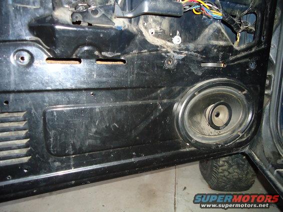 Inside of a vehicle door panel with a speaker and exposed wiring. A tire is visible below