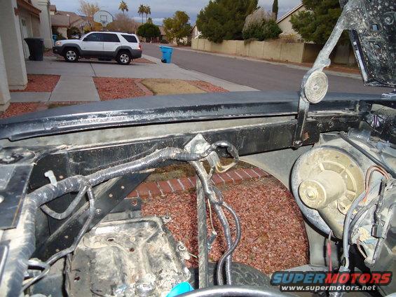 Engine bay of a dark-colored pickup truck, showing wiring and components. A white SUV is parked in the background