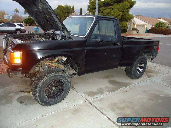 Black pickup truck, hood open, showing exposed engine components. Front fender removed, revealing wiring. Aftermarket wheels