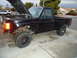 1987 Ford F-150 - A black pickup truck sits in a driveway with its hood up, revealing its engine compartment and exposed wiring.