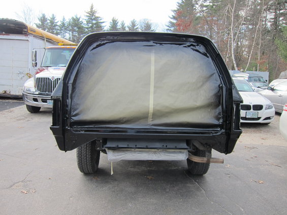 Rear view of a black pickup truck bed covered with plastic sheeting for painting. Other vehicles and a large truck are visible in the background