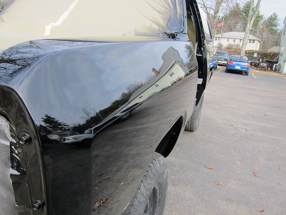 Close-up of a shiny black pickup truck fender and door, with reflections of houses and trees. Other vehicles are parked in the background